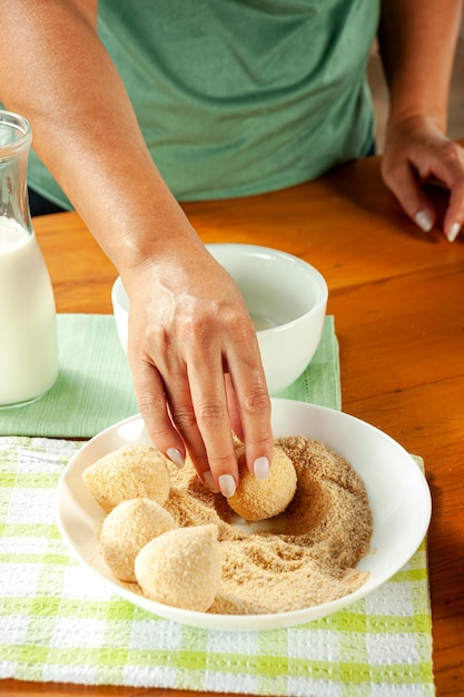 Woman hands breading brazilian croquette coxinha de frango with breadcrumbs on a wooden kitchen table