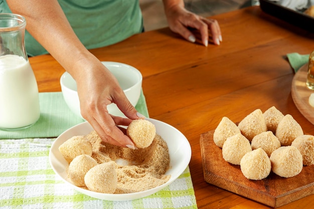 Woman hands breading brazilian croquette coxinha de frango with breadcrumbs on a wooden kitchen table