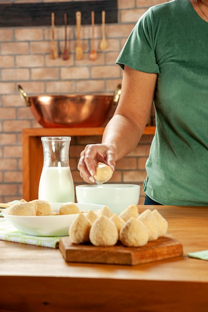 Woman hands breading brazilian croquette coxinha de frango with breadcrumbs on a wooden kitchen table