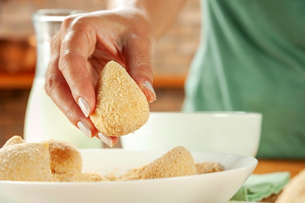 Woman hands breading brazilian croquette coxinha de frango with breadcrumbs on a wooden kitchen table