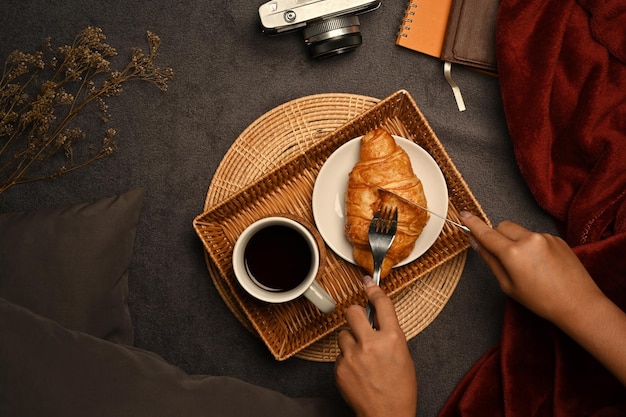 Woman hands cutting a piece of croissant by knife and fork for breakfast with coffee cup on wooden tray