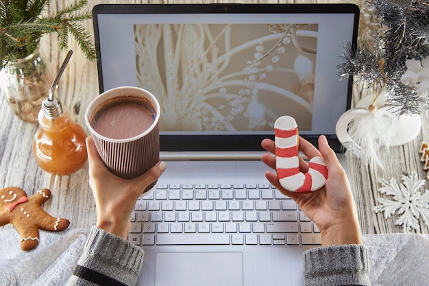 Woman hands hold a cardboard cup of cocoa and gingerbread Festive home office laptop mockup decorated Christmas tree Remote work watching webinar studying or calling family friends