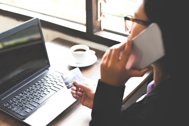 Woman hands holding credit card and using laptop and mobile phone for online shopping in a office table.