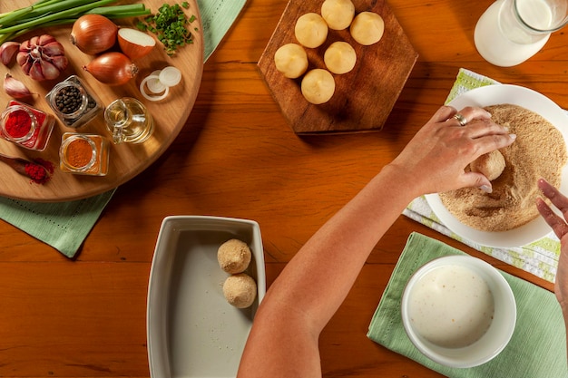 Woman hands preparing brazilian croquette coxinha de frango with breadcrumbs on a wooden kitchen table Top view