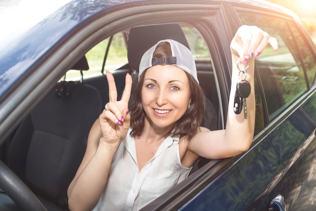 woman in a hat holding keys to new bought car and smiling at camera