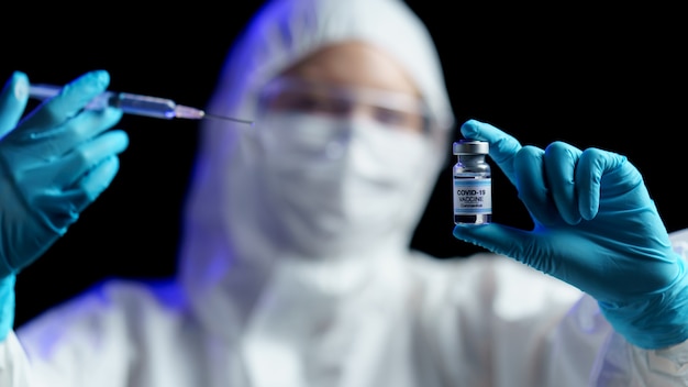 Woman in hazmat suit with vaccine and syringe injection for prevention, immunization and treatment from corona virus infection.