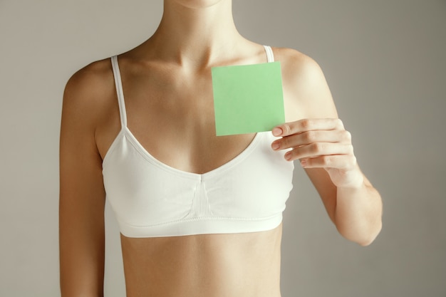 Woman health. Female model holding empty card near breast. Young adult girl with paper for sign or symbol isolated on gray studio background. Cut out part of body. Medical problem and solution.