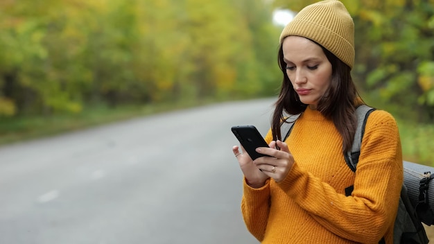 Woman hiker in orange sweater walks on empty road and types message to friend on phone