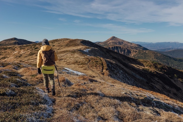 Woman hiker in yellow jacket with backpack climbs autumn mountains with light snow on ground on sunn