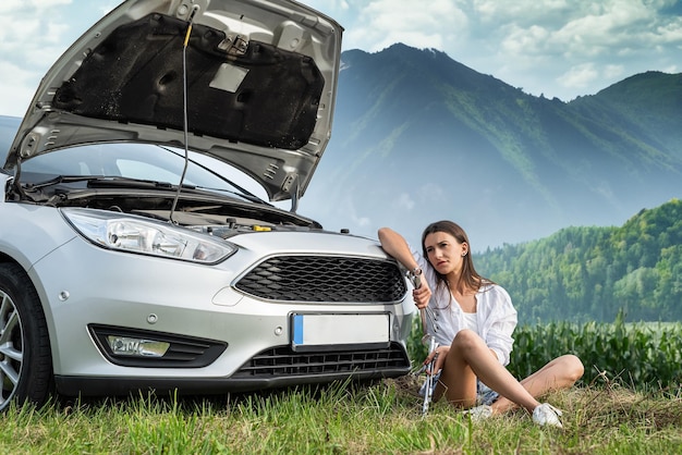 Woman hold a wrench and stand near her broken car with the open hood for repairs at road