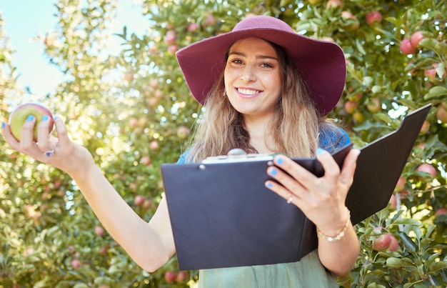 Woman holding an apple and a clipboard on a sustainable orchard farm in summer Happy farmer doing stock taking and checking a checklist while harvesting fresh organic green and red fruit from trees