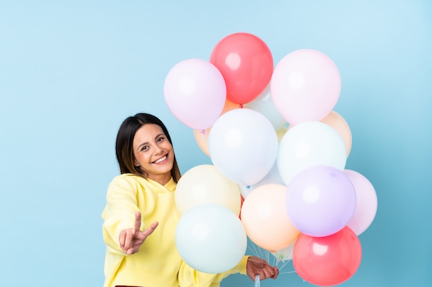 Woman holding balloons in a party over blue wall smiling and showing victory sign