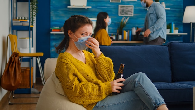 Woman holding beer bottle drinking looking at camera, relaxing on sofa, spending time with friends keeping social distancing with face mask in living room preventing spread of coronavirus.