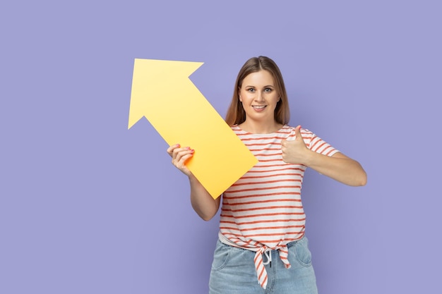 Woman holding big yellow arrow pointing aside and showing thumb up like gesture