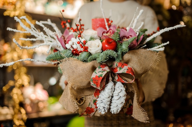 woman holding a christmas composition with pink orchids, white roses, fir-tree branches, red apple and candle in the sackcloth in the flower shop