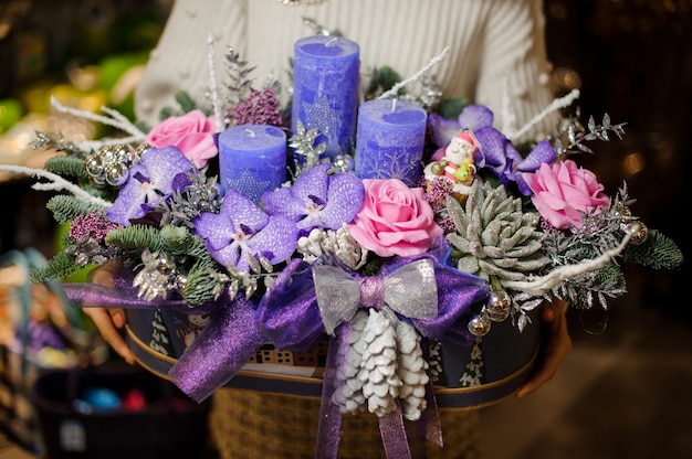 woman holding a christmas composition with purple and pink flowers, succulents, fir-tree branches and candles in the box