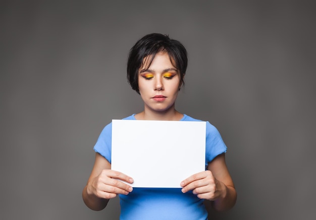 A woman holding an empty card a banner made of white paper isolated on a gray wall Portrait of an unhappy sad young woman Human facial expressions emotions Advertising concept