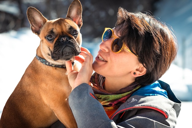 Woman holding on hands French buldog and playing with it on the white snowy background