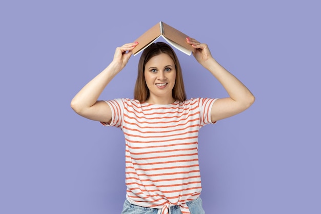 Woman holding opened book on head looking at camera with smile expressing positive emotions