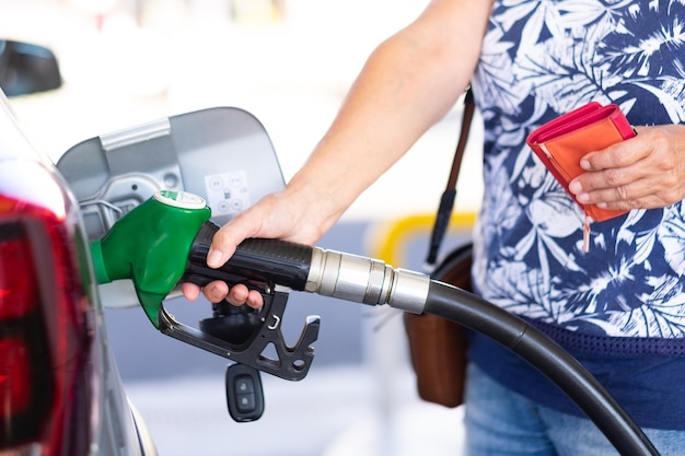 Woman holding petrol gasoline fuel pump to refuel her silver car at the service station, holding wallet to pay