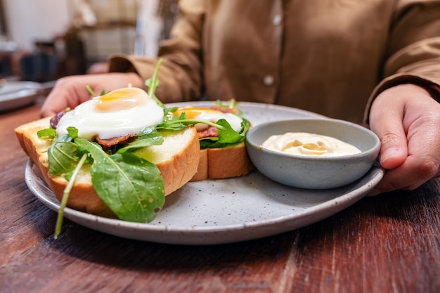 A woman holding a plate of breakfast sandwich with eggs, bacon and sour cream on wooden table