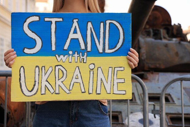 Photo woman holding poster in colors of national flag with words stand with ukraine near broken tank on city street closeup