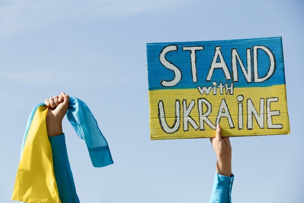 Photo woman holding poster stand with ukraine and national flag against blue sky closeup