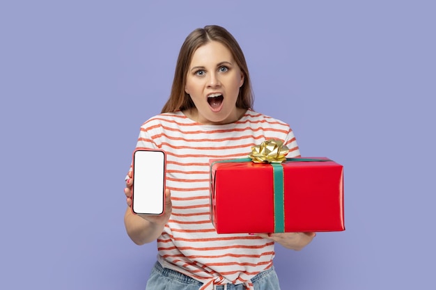 Woman holding present box and showing cell phone with white screen with copy space for advertisement