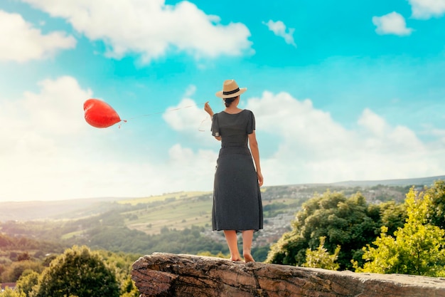 Woman holding a red balloon in the shape of a heart on top of the hill against the blue cloudy sky