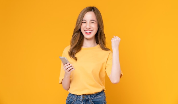 woman holding smartphone with fists clenched celebrating victory expressing success on light orange