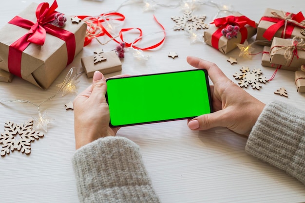 Woman holding smartphone with green screen on white wooden table background with Christmas gifts