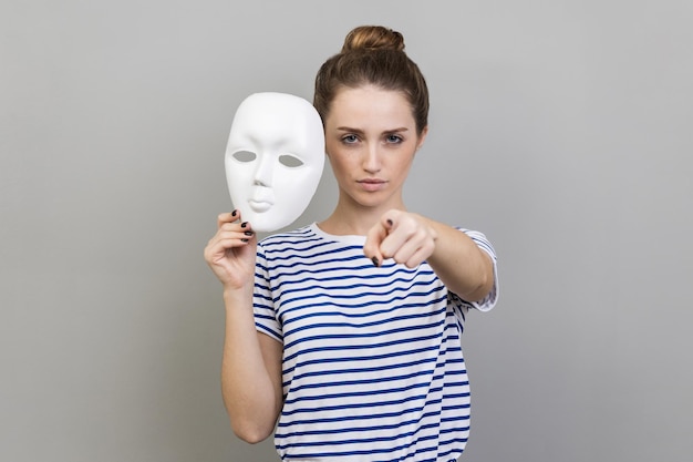 Woman holding white mask in hand pointing finger to camera with serious expression