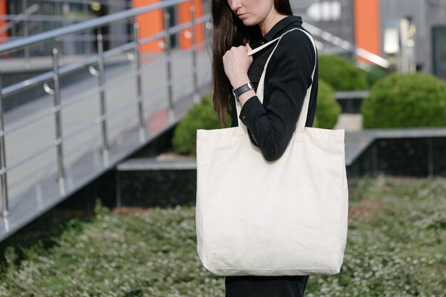 Woman holding white textile eco bag against urban city background Ecology or environment protection concept White eco bag for mock up