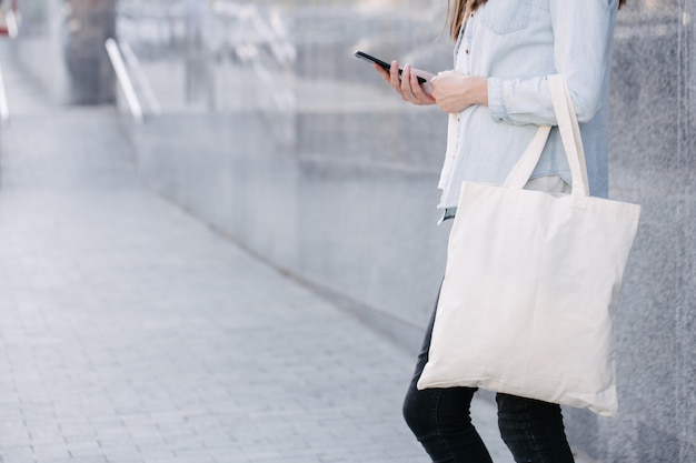 Woman holding white textile eco bag against urban city background. . Ecology or environment protection concept. White eco bag for mock up.