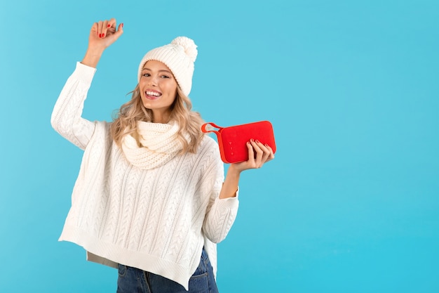 woman holding wireless speaker listening to music happy dancing wearing white sweater and knitted hat on blue