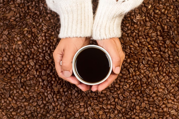 Woman holding with both hands a cup of black coffee on a heap of coffee grains in a top view