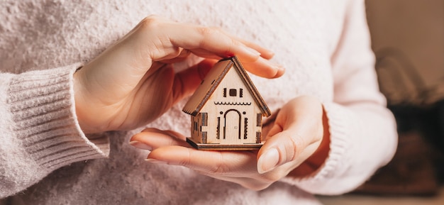 Woman holding a wooden house with her hands with the sun on a light pink background. Sweet home