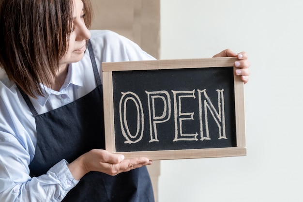 Woman Holds Board with Text Open.  Worker in Apron Shows Opening of the Cafe or Market.