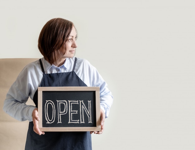 Woman Holds Board with Text Open.  Worker in Apron Shows Opening of the Cafe or Market.