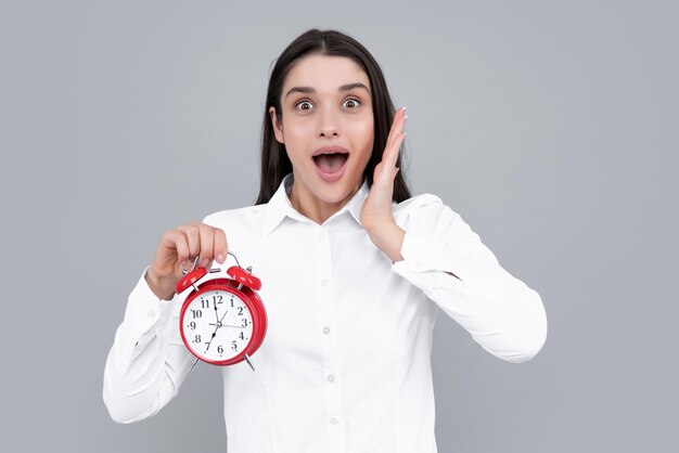 Woman holds a clock Emotional young female face Portrait of young surprised woman with alarm clock on grey background