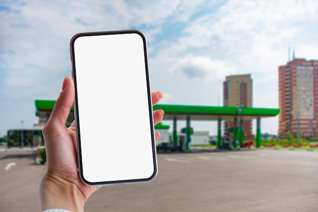 Woman holds a closeup of a smartphone with a white screen in his hands against the backdrop on a gas station Technology mockup for apps and websites