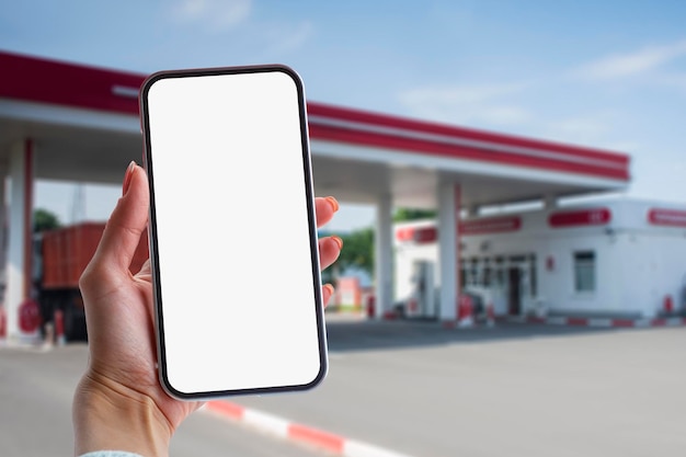 Woman holds a closeup of a smartphone with a white screen in his hands against the backdrop on a gas station Technology mockup for apps and websites