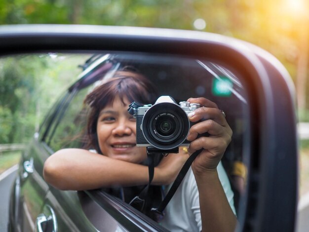 A woman holds a digital camera and takes a picture of herself smiling reflected in the car mirror