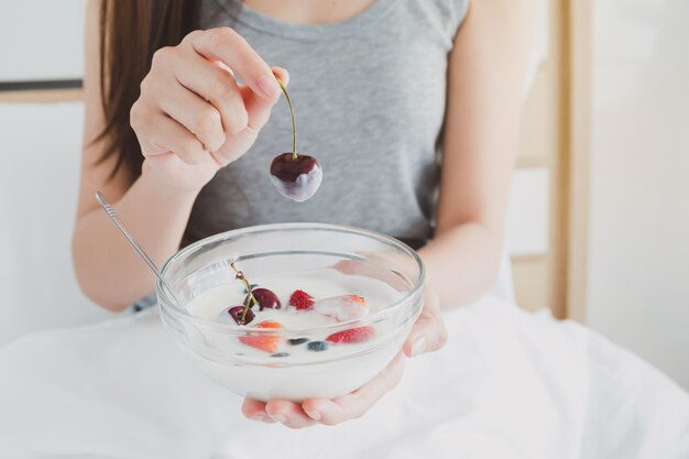 The woman holds a glass bowl of yogurt and strawberry fruit on white bed in morning.