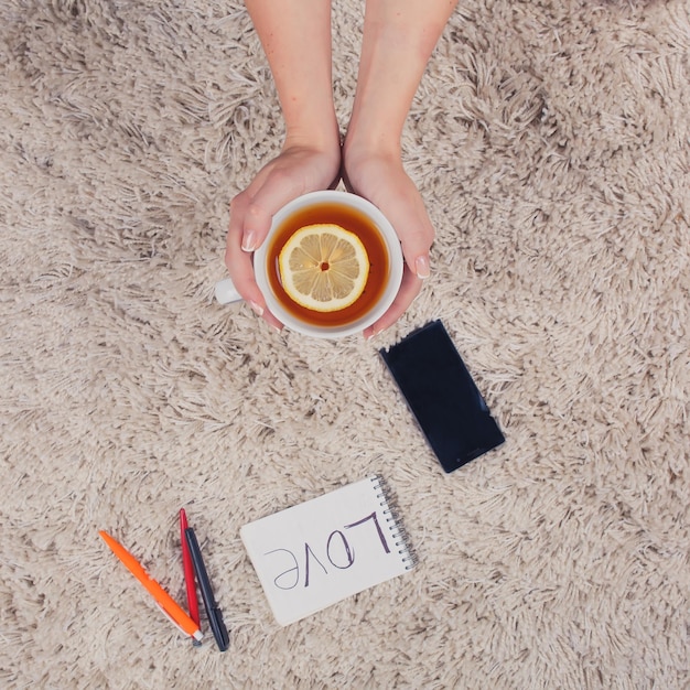 A woman holds hands a cup of tea with lemon phone pen and notebook lying on the carpet top view