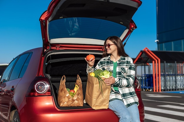 Woman holds orange pumpkin and shopping paper bag with a groceries sitting on cars trunk