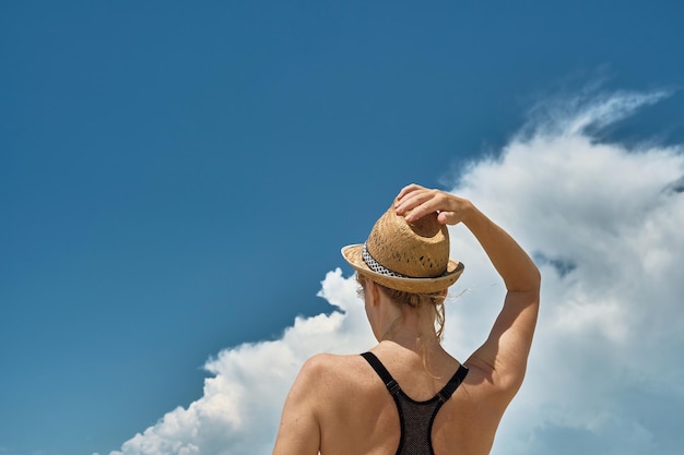 Woman holds a straw hat on her head with her hand a silhouette of a beautiful blonde against the background of cumulus clouds over the sea