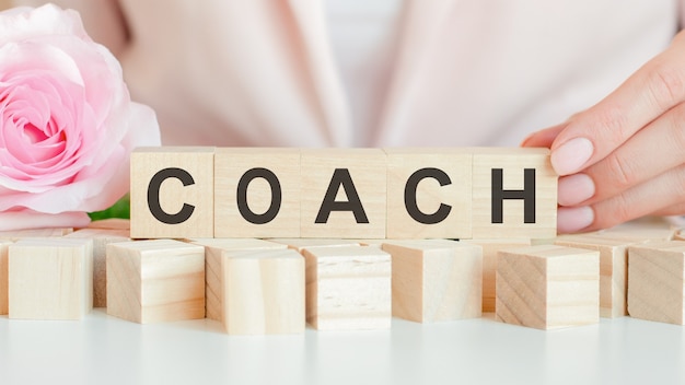 Woman holds a wooden cube with the text of coach in her hand. on the wooden cubes there is a living rose flower. pink background, front view. business, economic, education concept