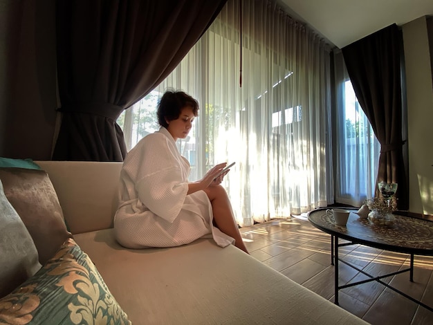 Woman in home or apartment Girl in white bathrobe stands in front of window with smartphone