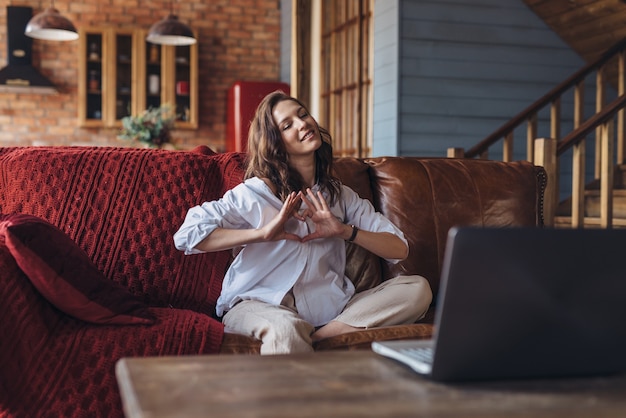 Woman at home speaks to a loved one on video call and shows heart gesture as symbol of love.